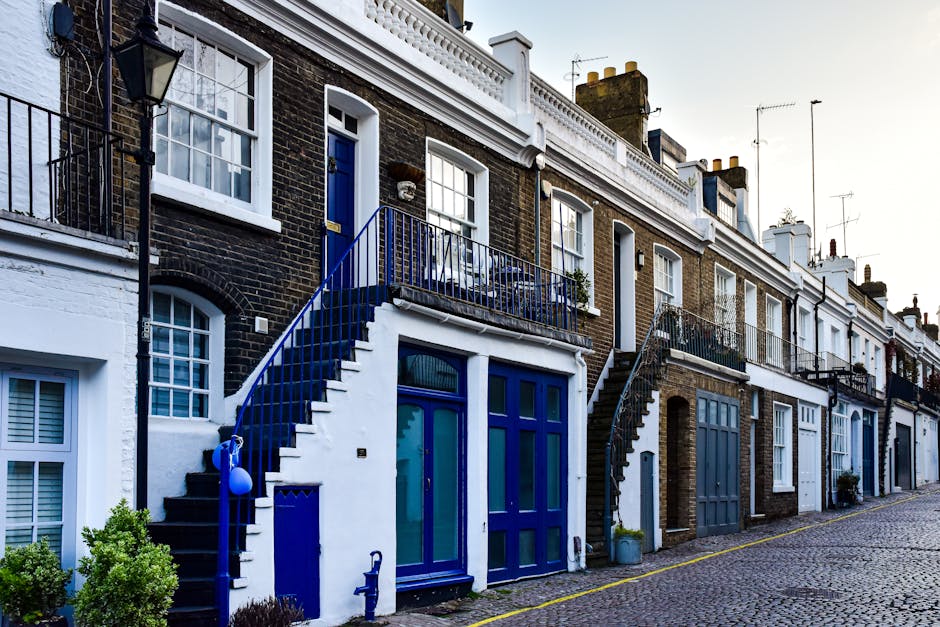 The image shows a row of terraced houses with brick exteriors, each featuring small front gardens and external staircases leading to upper-level entrances. The house in the foreground has a white-pain