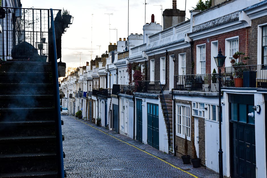 The image shows a row of terraced houses with brick exteriors, each featuring small front gardens and external staircases leading to upper-level entrances. The house in the foreground has a white-pain