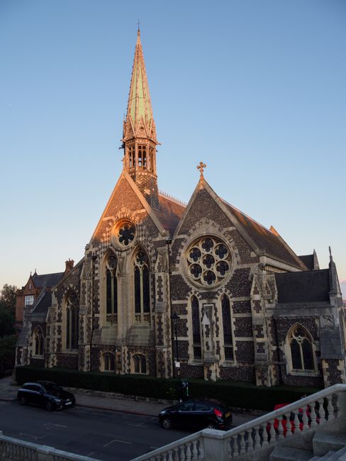 A large historic church building with intricate Gothic architectural features, including pointed arches, decorative stone tracery, and circular stained glass windows. The church's exterior is constructed from patterned stonework with a combination of light and dark materials, creating a contrasting effect. The building has a tall, slender spire topped with a cross, rising above the main structure. The church is situated near a street with parked cars visible at the roadside, and a low hedge runs along the base of the building. The setting appears to be during the late afternoon or early evening, with warm sunlight illuminating the upper parts of the spire and facade, highlighting the detailed craftsmanship. This image relates to house removals and relocation services, capturing a notable local landmark that could be part of a community or heritage area in Ealing, as referenced in the Ealing Broadway moving advice and local removals guide provided by Man with Van Ealing.