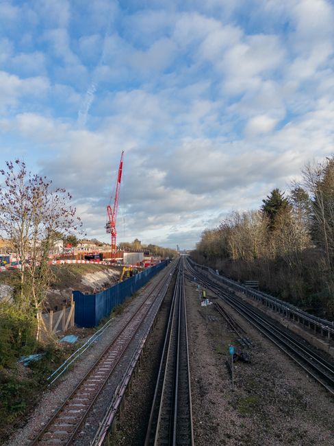A view of multiple railway tracks extending into the distance under a partly cloudy sky, with a construction site on the left side featuring a red crane and blue fencing along the perimeter. On the right side, there are trees without leaves, indicating a possible autumn or winter season. The scene is outdoors, with no people visible, and shows the space where home relocation and furniture transport might occur during a house move. The railway and construction elements align with logistical aspects of moving services provided by Man with Van Ealing, as part of the site's guidance on local removals and moving advice in Ealing.