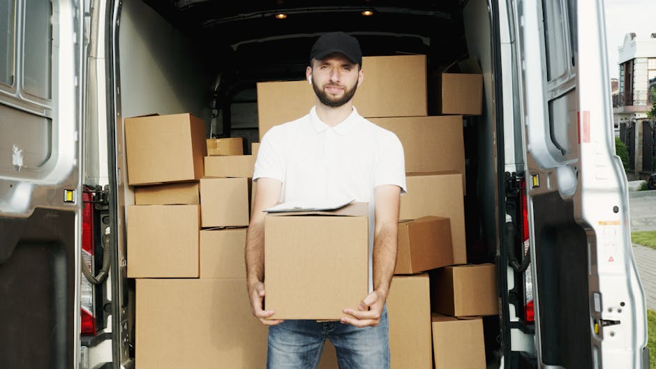 A man with a beard, wearing a white polo shirt and a black cap, is standing in front of an open moving van during a home relocation. He is holding a packed cardboard box with both hands, positioned in front of his torso. The interior of the van is filled with multiple cardboard boxes of various sizes, some stacked on top of each other, and secured with plastic wrapping and packing materials. The van is parked outside a residential property, with a paved driveway and nearby houses visible in the background. The scene depicts the loading process involved in furniture transport and packing for a house move, with lighting indicating daytime. The image illustrates the services offered by Man with Van Ealing in residential removals, focusing on packing and moving logistics.