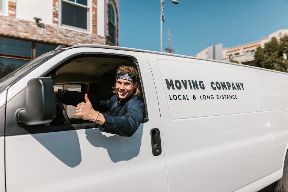 A young male mover inside a white movement van with 'MOVING COMPANY LOCAL & LONG DISTANCE' printed on the side, smiling and giving a thumbs-up through the open driver’s window. The van is parked on a city street during daylight with modern buildings, a clear blue sky, and utility poles visible in the background. The driver appears to be wearing a dark jacket and a bandana on his head, indicating a professional home relocation service. This scene captures the loading process for furniture transport and packing during a house move, with the focus on ensuring safe and efficient handling of belongings, as facilitated by Man with Van Ealing. The image emphasizes the importance of proper vehicle use and positive service attitude in house removals within Ealing.